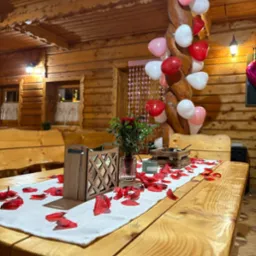 Dekoracja stołu z drewnianymi elementami, płatkami róż i balonami w rustykalnym wnętrzu. Romantyczna aranżacja na specjalną okazję, ciepłe oświetlenie.