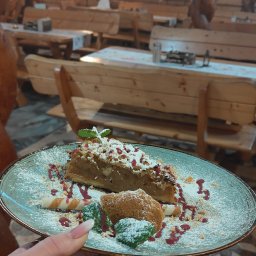 AKK SPÓŁKA Z OGRANICZONĄ ODPOWIEDZIALNOŚCIĄ - Kawałek szarlotki na talerzu, posypany cukrem pudrem i udekorowany sosem malinowym i miętą, trzymany w dłoni na tle drewnianej restauracji.