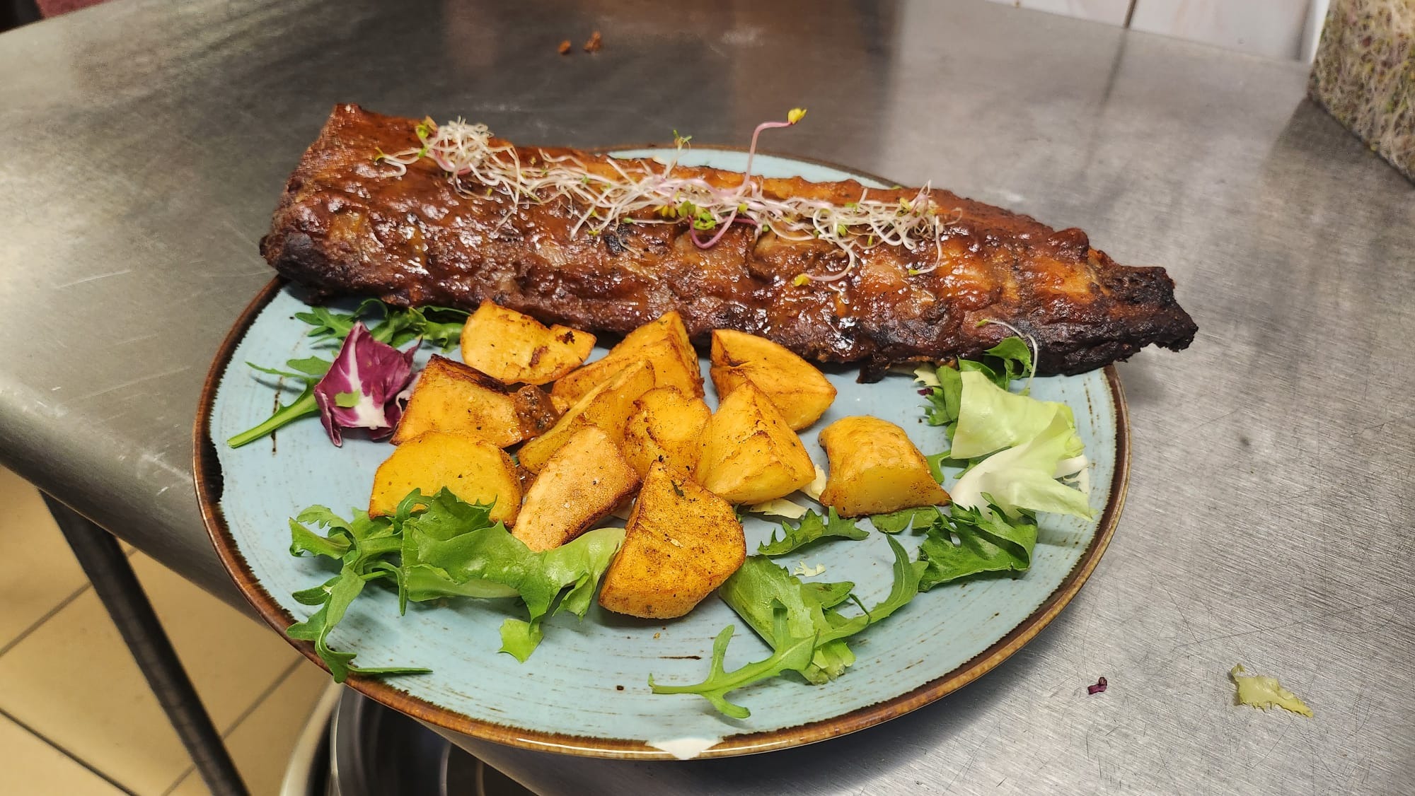 Żeberka BBQ z ziemniakami i zielenią na turkusowym talerzu z brązową obwódką, na metalowym blacie. Żeberka udekorowane kiełkami.