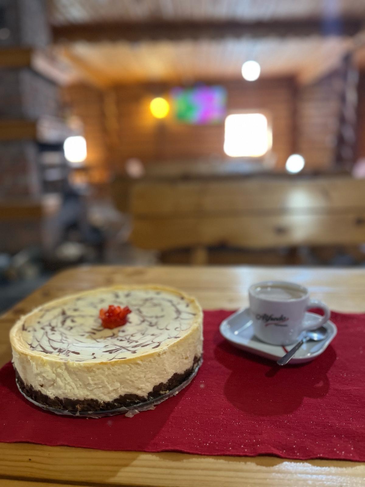 Okrągły sernik z dekoracją z truskawek i filiżanka kawy na czerwonej serwetce, na drewnianym stole. W tle drewniana konstrukcja budynku i rozmyte światła.