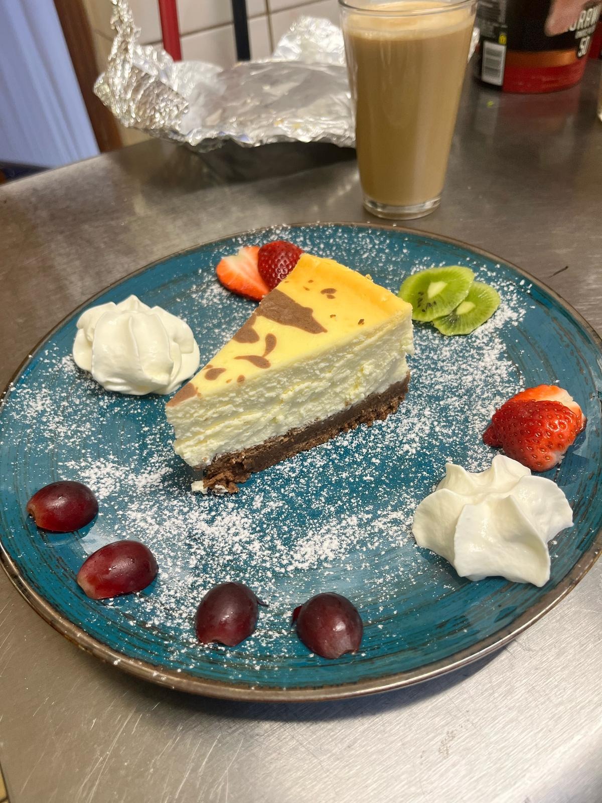 Turkusowy talerz z sernikiem, bitą śmietaną, truskawkami, kiwi i winogronami, oprószony cukrem pudrem. W tle napój w szklance i opakowania produktów.