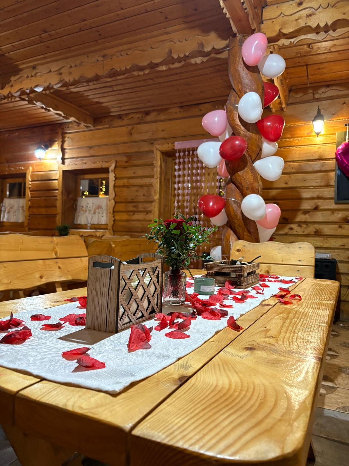 Dekoracja stołu z drewnianymi elementami, płatkami róż i balonami w rustykalnym wnętrzu. Romantyczna aranżacja na specjalną okazję, ciepłe oświetlenie.