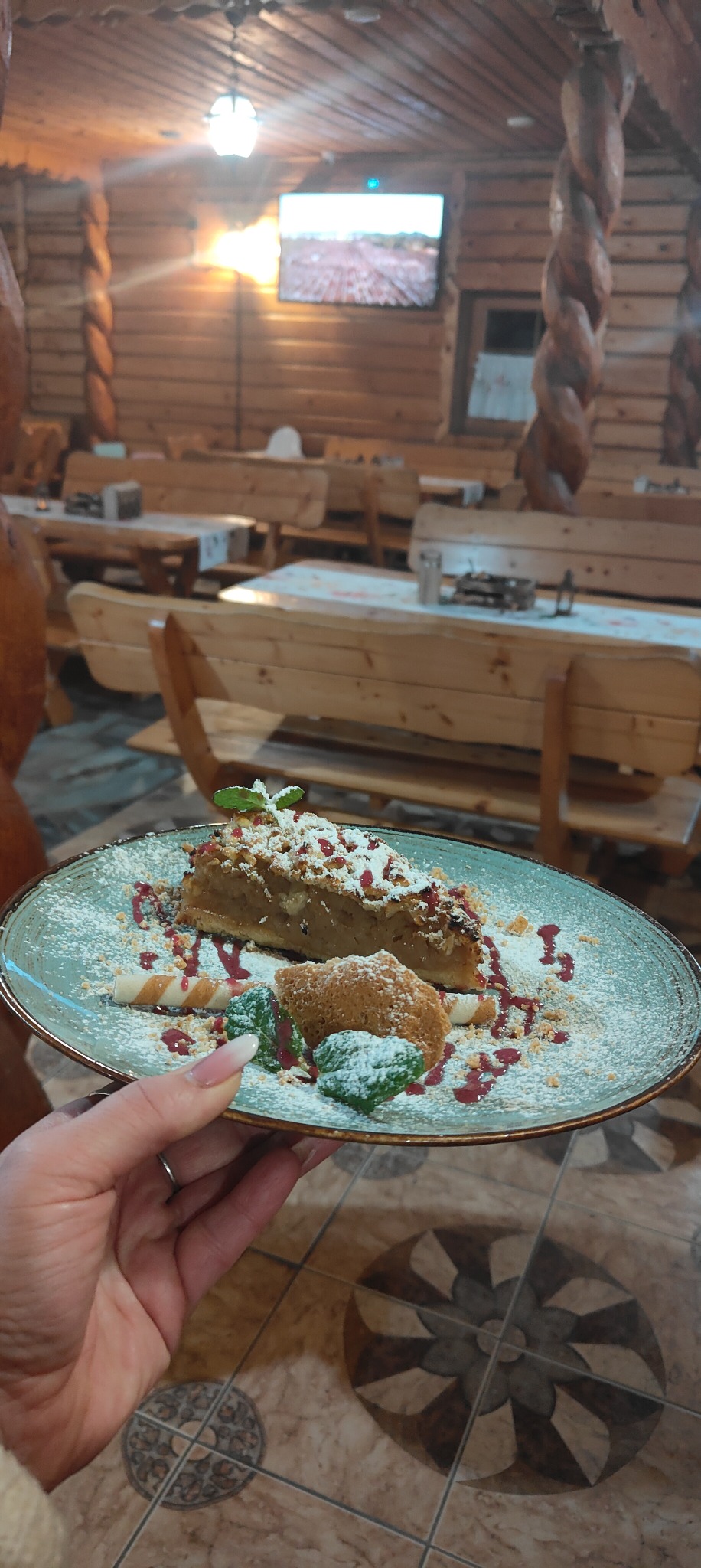 Kawałek szarlotki na talerzu, posypany cukrem pudrem i udekorowany sosem malinowym i miętą, trzymany w dłoni na tle drewnianej restauracji.