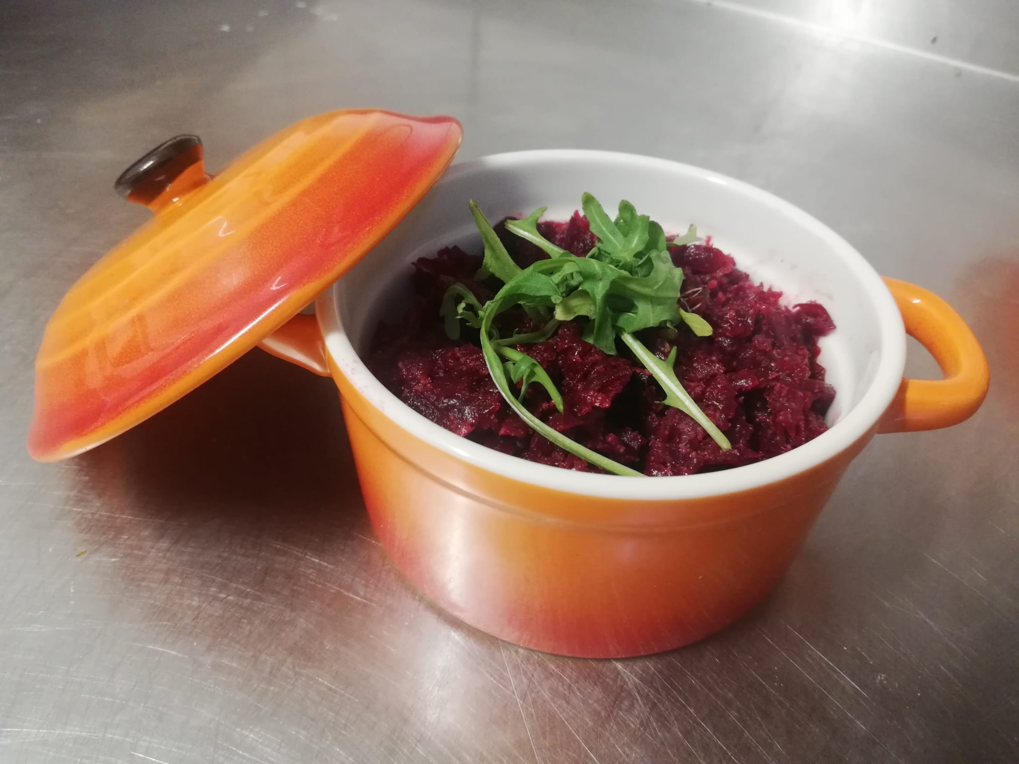 Buraczki tarte w pomarańczowej miseczce z rukolą na metalowym blacie. Pokrywka obok. Danie gotowe do serwowania, catering.