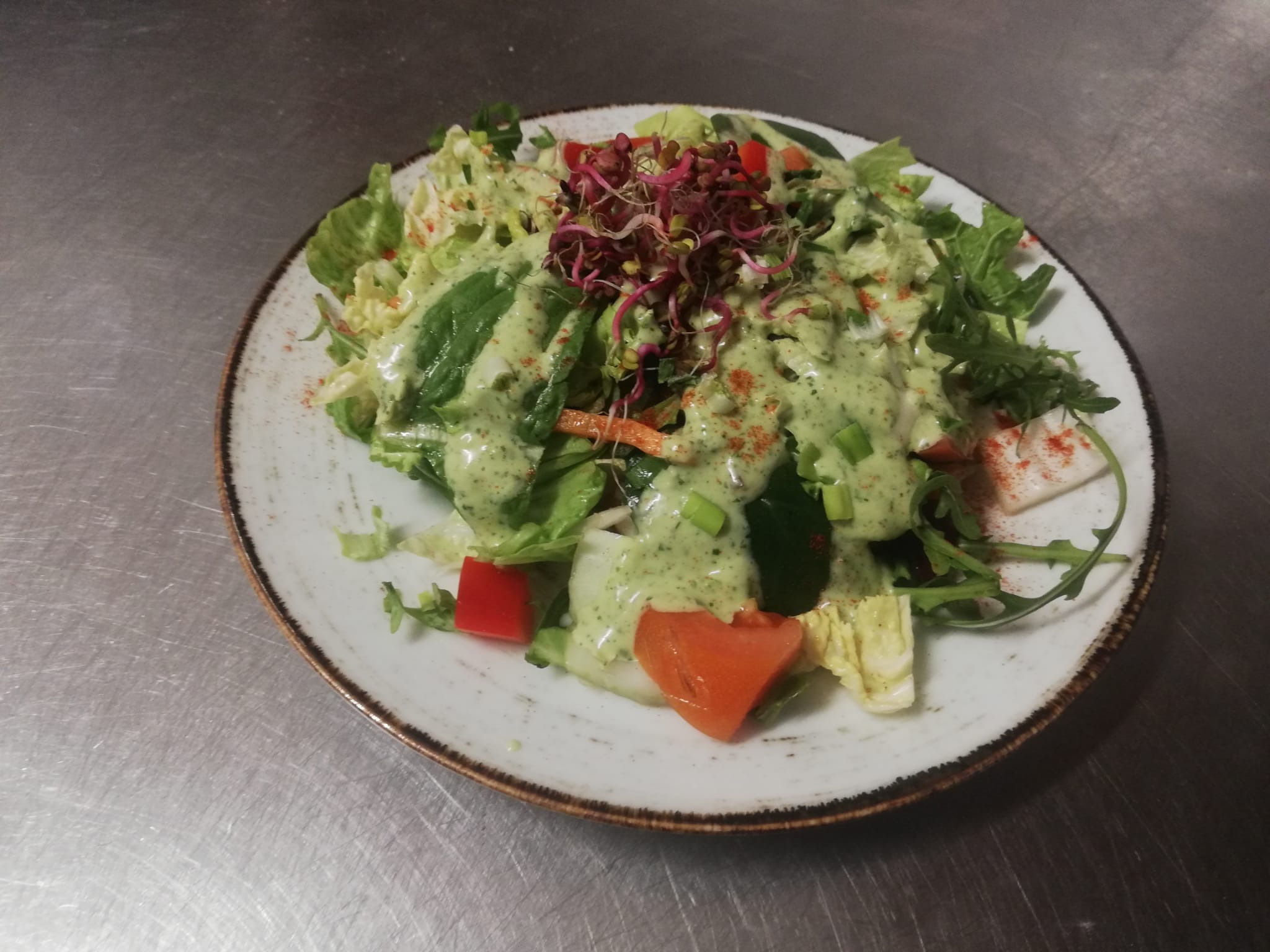 Świeża sałatka z warzywami, polana kremowym sosem i posypana czerwoną papryką, podana na talerzu z wzorem, na metalowym tle.