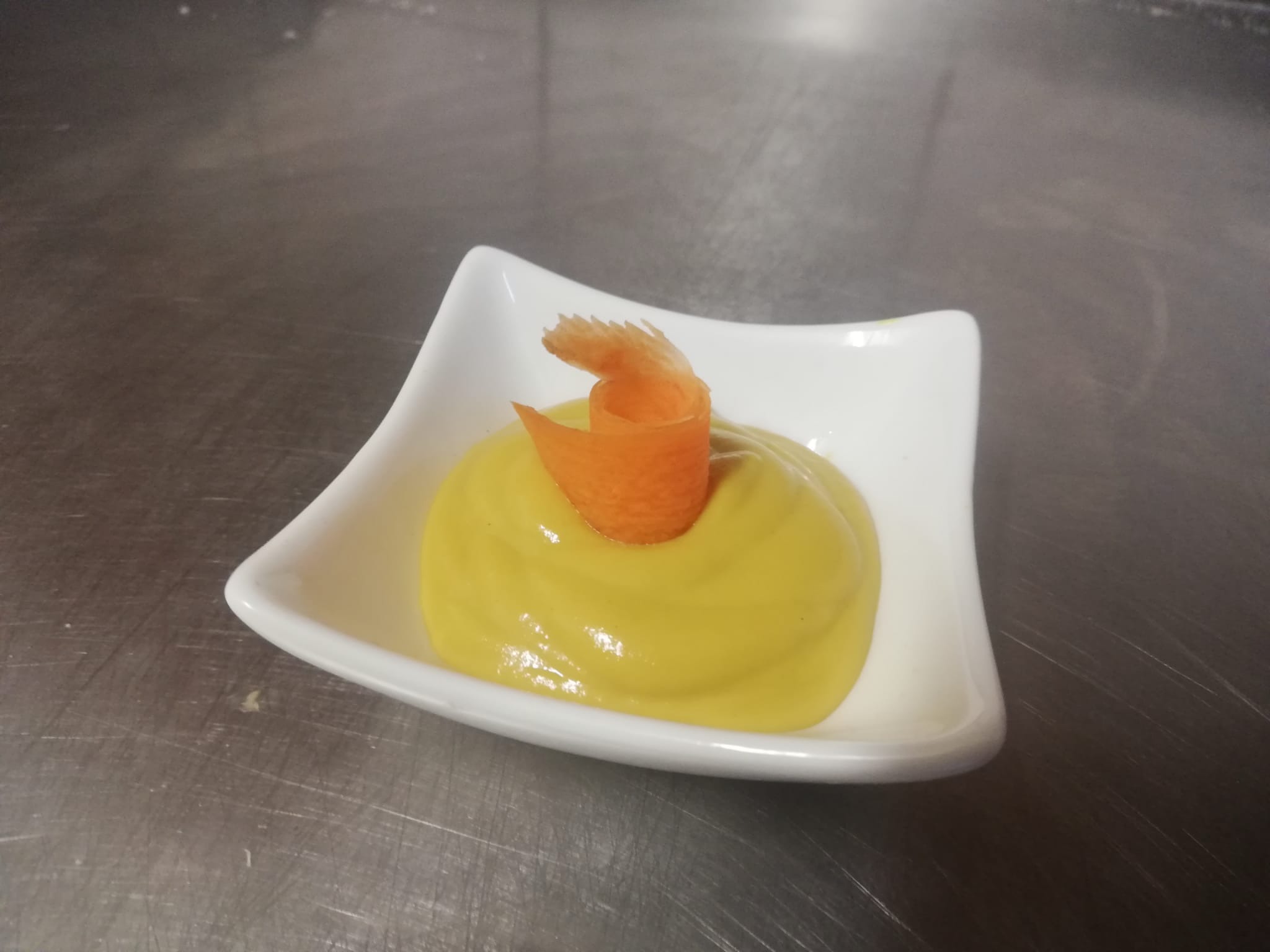 Elegancko podane danie: kremowy sos w białej miseczce z dekoracją z marchewki, na metalowym blacie. Finezyjne detale prezentacji kulinarnej.