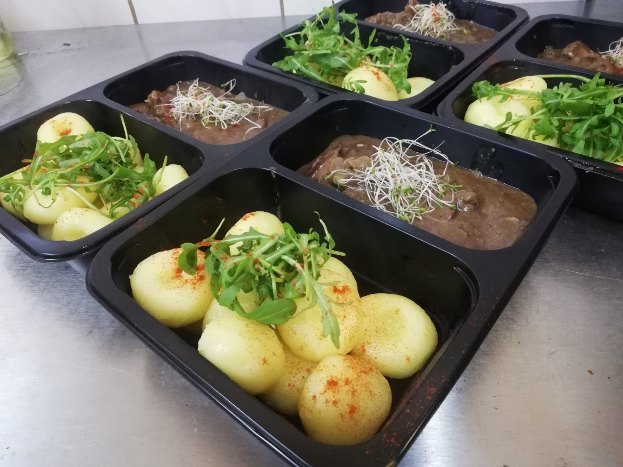 Catering: Porcje w czarnych pojemnikach z ziemniakami posypanymi papryką, sosem i zieleniną. Gotowe dania na metalowym blacie, przygotowane do transportu.