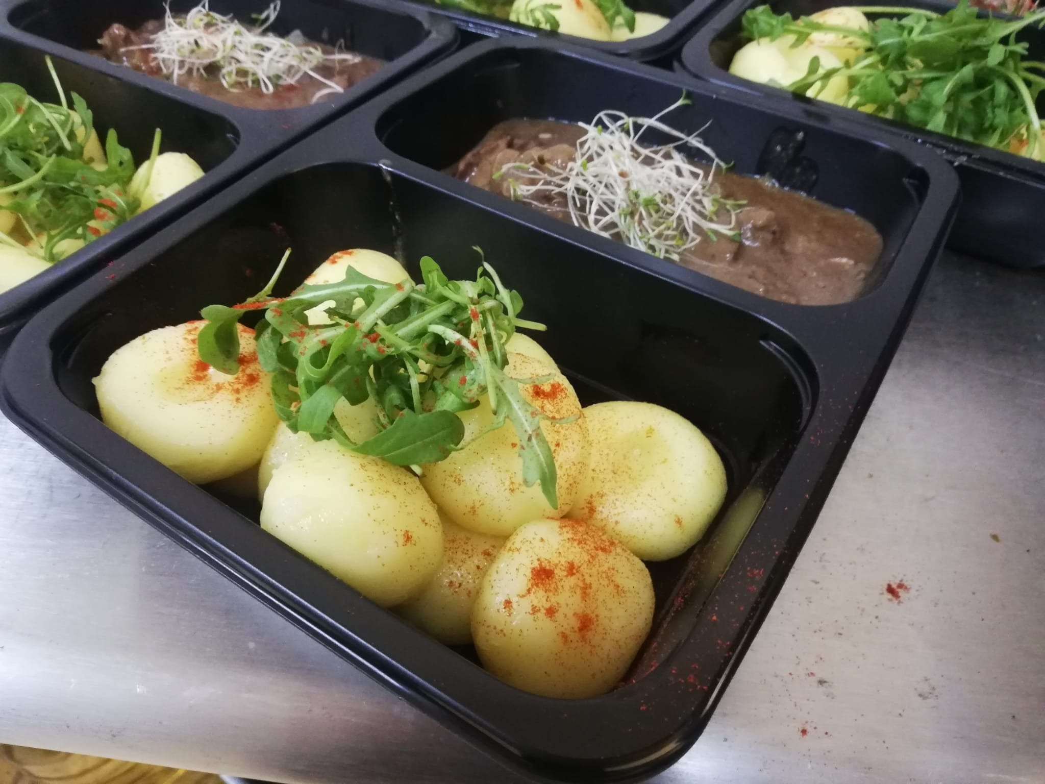 Porcje cateringowe w czarnych pojemnikach: ziemniaki z papryką i rukolą, sos mięsny z kiełkami. Widok z góry, ostre światło, metalowe tło. Gotowe dania.