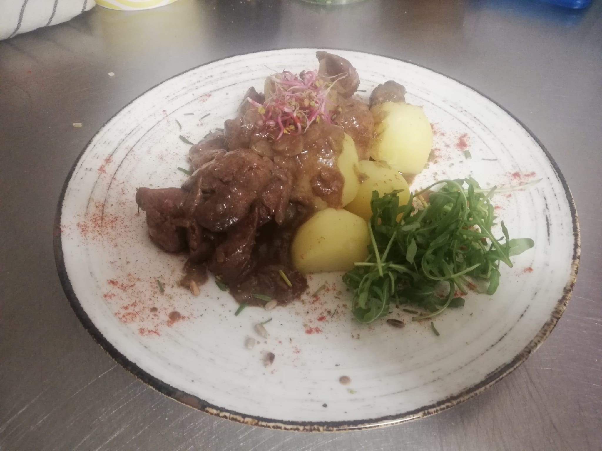 Danie na talerzu: gulasz wołowy z ziemniakami i rukolą, udekorowane kiełkami i przyprawami. Ceramiczny talerz z ciemnym obramowaniem, widoczne ziarna słonecznika.