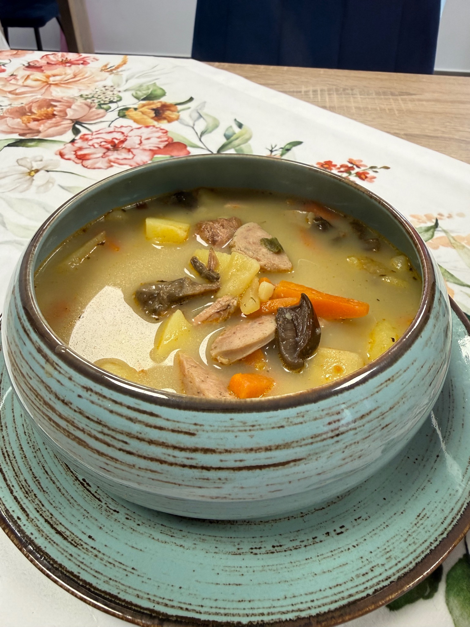 Zupa w turkusowej misce na talerzu, z ziemniakami, marchewką i grzybami, na stole z kwiatowym obrusem. Apetyczne danie gotowe do podania.