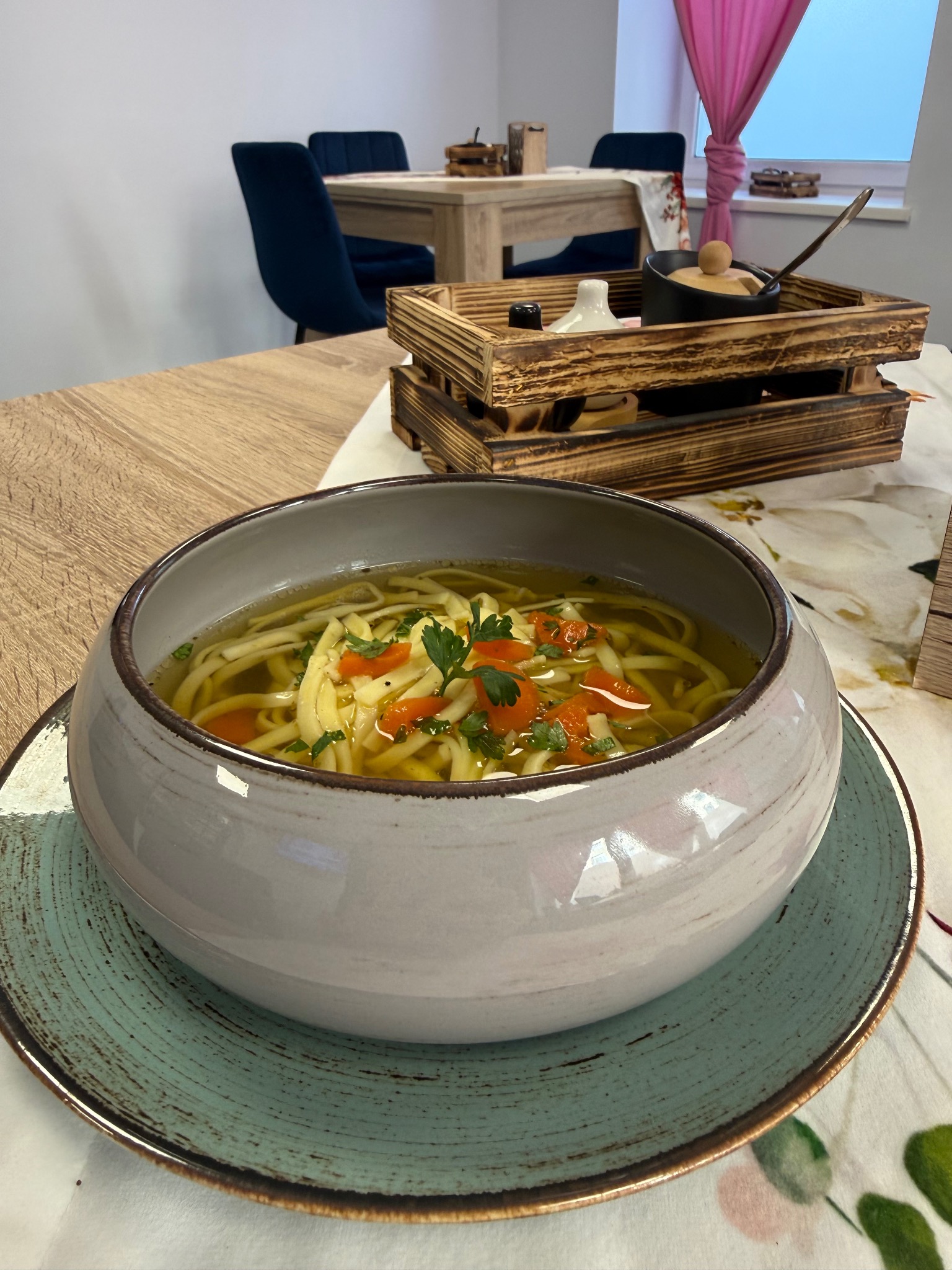 Aromatyczny rosół z makaronem i marchewką w ceramicznej misce na talerzu, w tle drewniana skrzynka z przyprawami i zastawą. Przytulna aranżacja stołu.