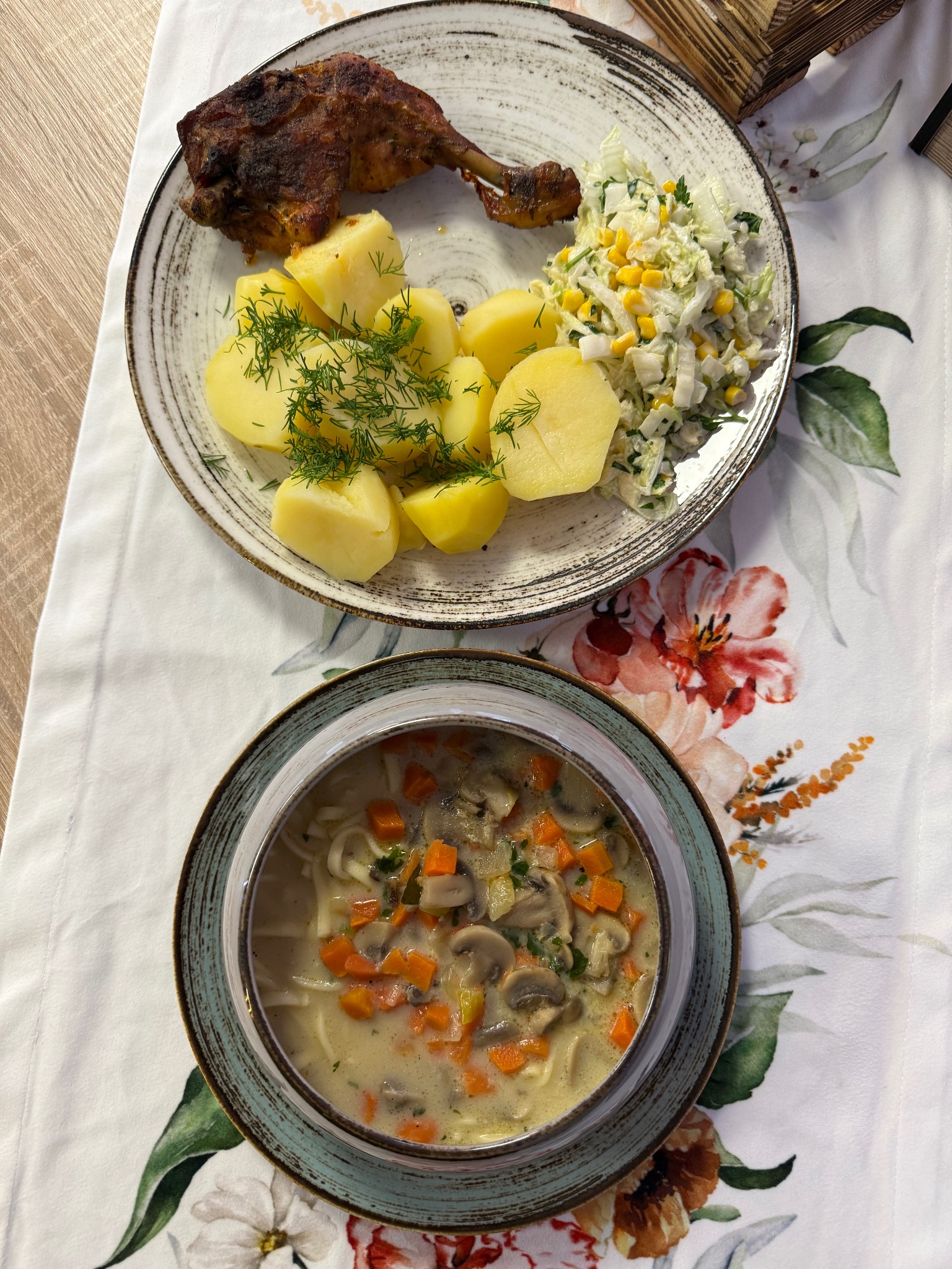 Zupa krem z grzybami i marchewką w misce oraz talerz z pieczoną nogą, ziemniakami z koperkiem i surówką na kwiecistym obrusie, ujęcie z góry.