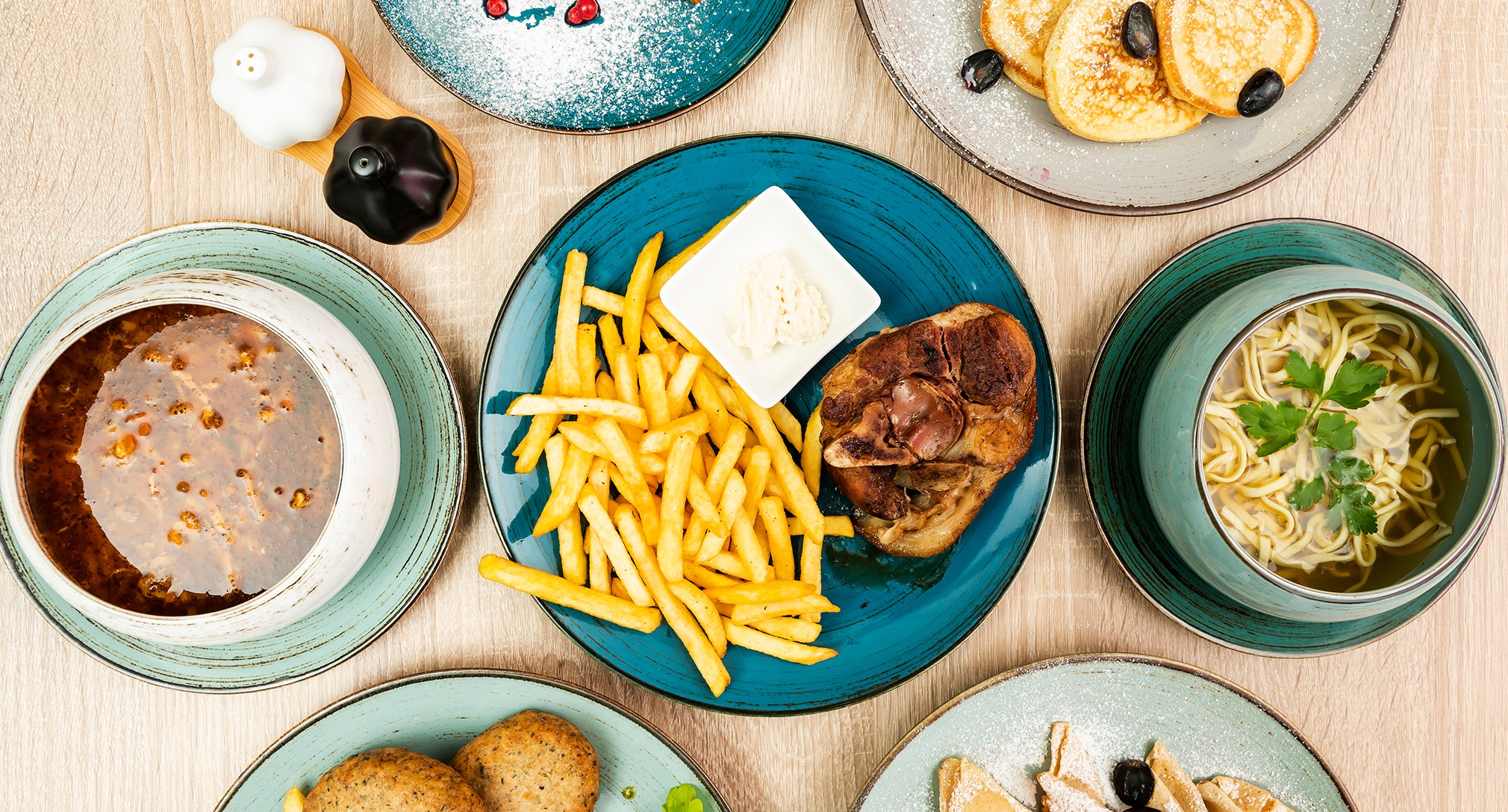 Kompozycja z różnorodnych dań: zupa, kotlety, naleśniki i frytki z sosem, podane na talerzach w odcieniach błękitu i zieleni, widok z góry.