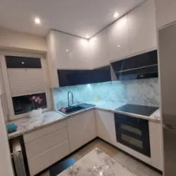 Biała kuchnia z marmurowym blatem i czarnymi akcentami. Widoczne okno z roletami i kwiatami. Nowoczesne wykończenie, oświetlenie punktowe.