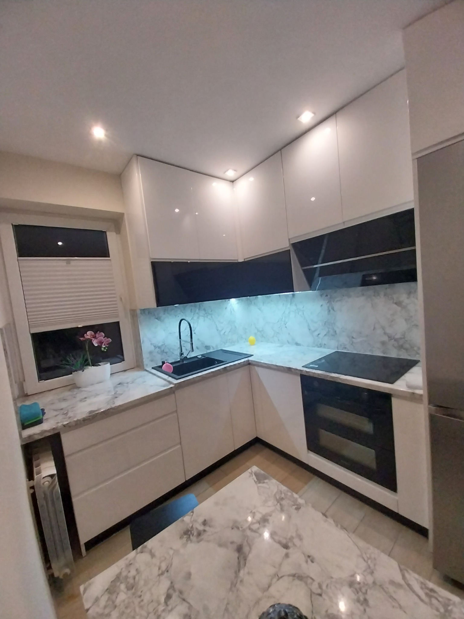 Biała kuchnia z marmurowym blatem i czarnymi akcentami. Widoczne okno z roletami i kwiatami. Nowoczesne wykończenie, oświetlenie punktowe.