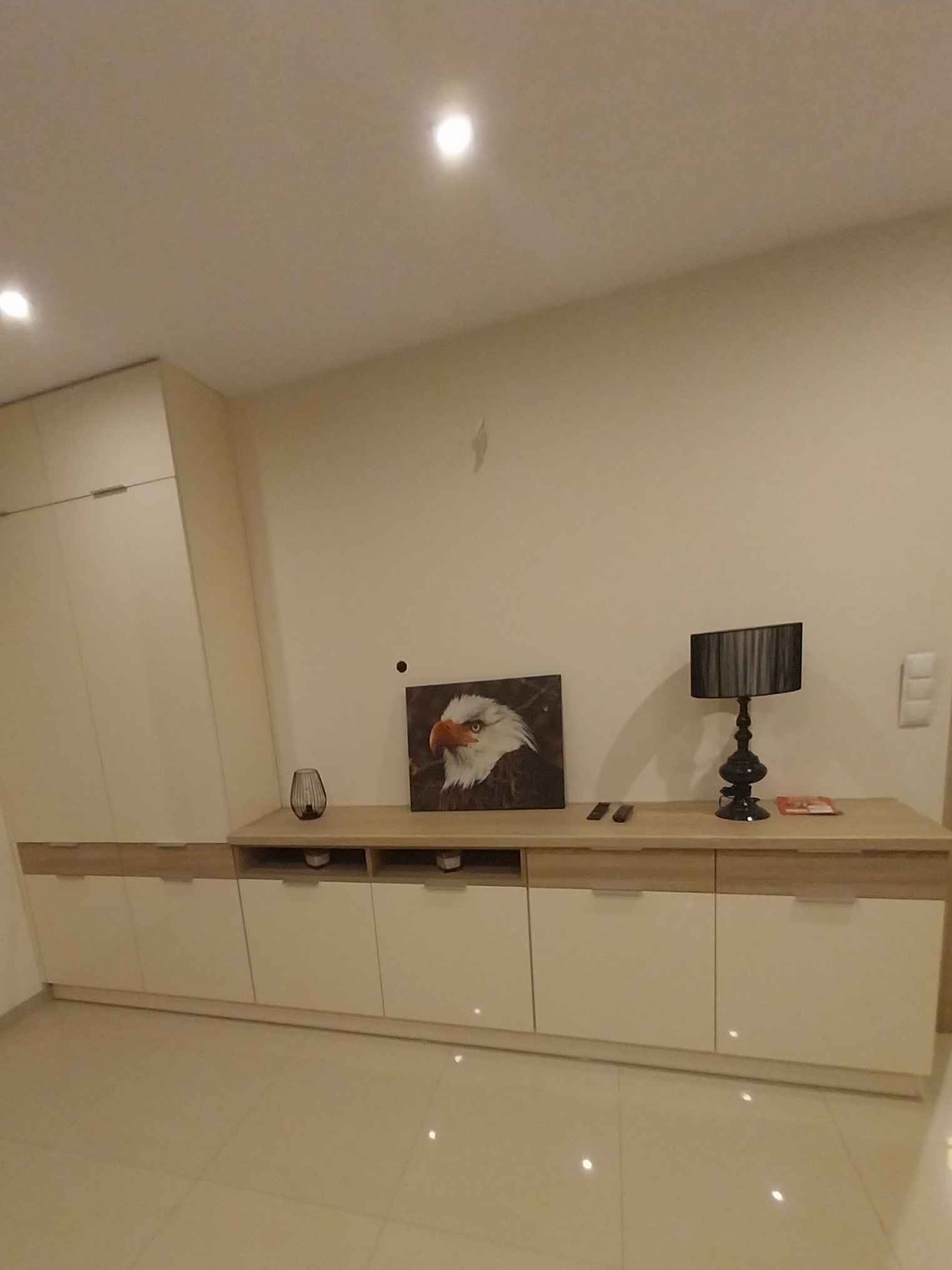 Jasna szafka RTV z obrazem orła i lampką. Fronty szafek bez uchwytów, na wysoki połysk. Całość w ciepłej tonacji, z widocznym fragmentem podłogi.