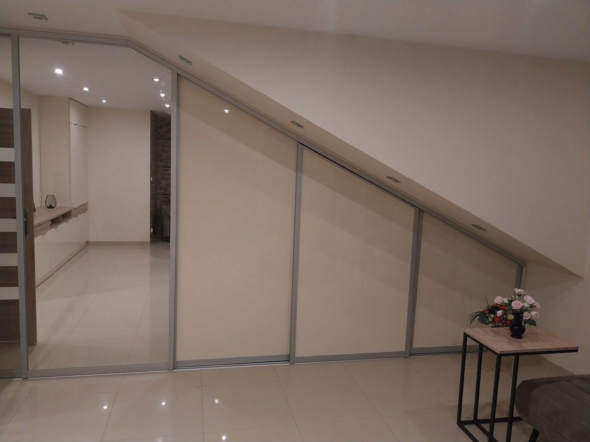 Beżowa szafa wnękowa na poddaszu ze szklanymi drzwiami przesuwnymi, odbijającymi wnętrze pokoju. Na pierwszym planie stolik z kwiatami.