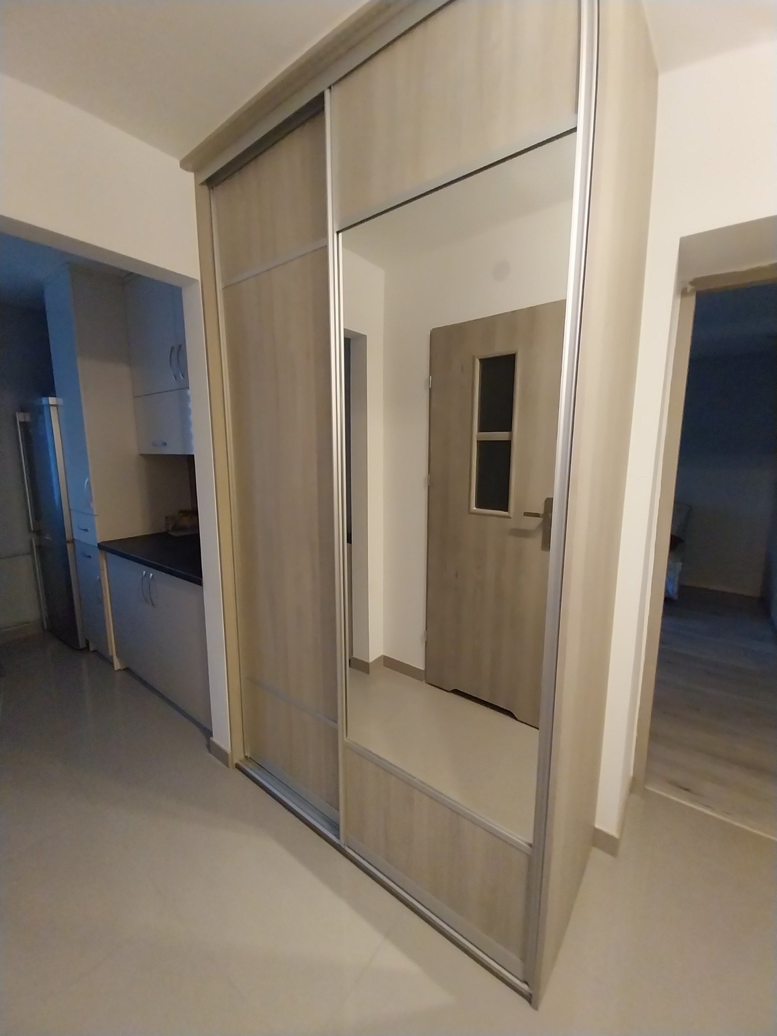 Jasna szafa przesuwna z lustrem w przedpokoju, odbijająca drzwi wejściowe. Widoczna część kuchni w tle, neutralna kolorystyka ścian i podłogi.