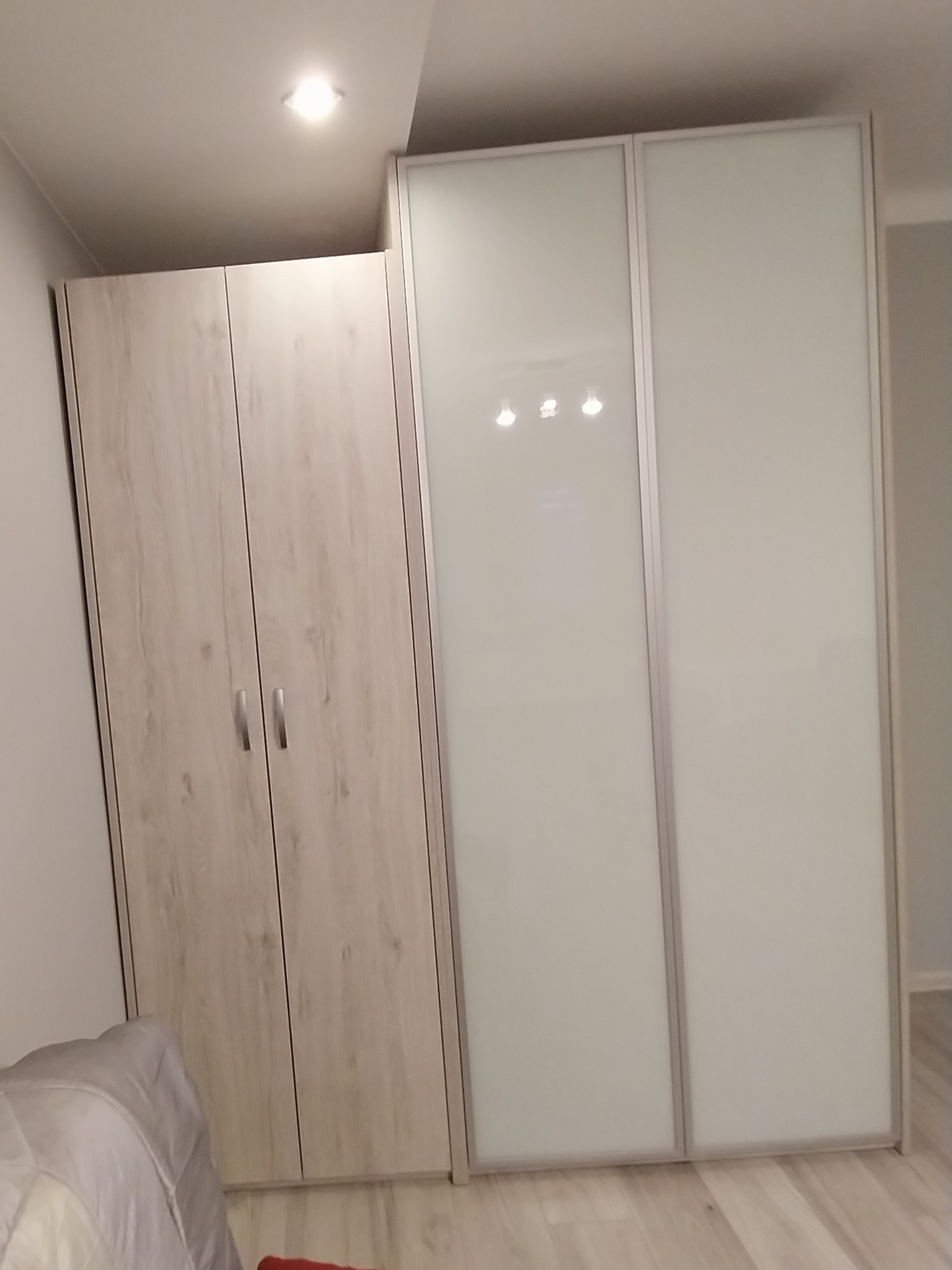 Nowoczesna szafa z dwojakimi drzwiami: drewnopodobne, dwuskrzydłowe i przesuwne z mlecznego szkła, w jasnym wnętrzu, oświetlona górnym światłem.