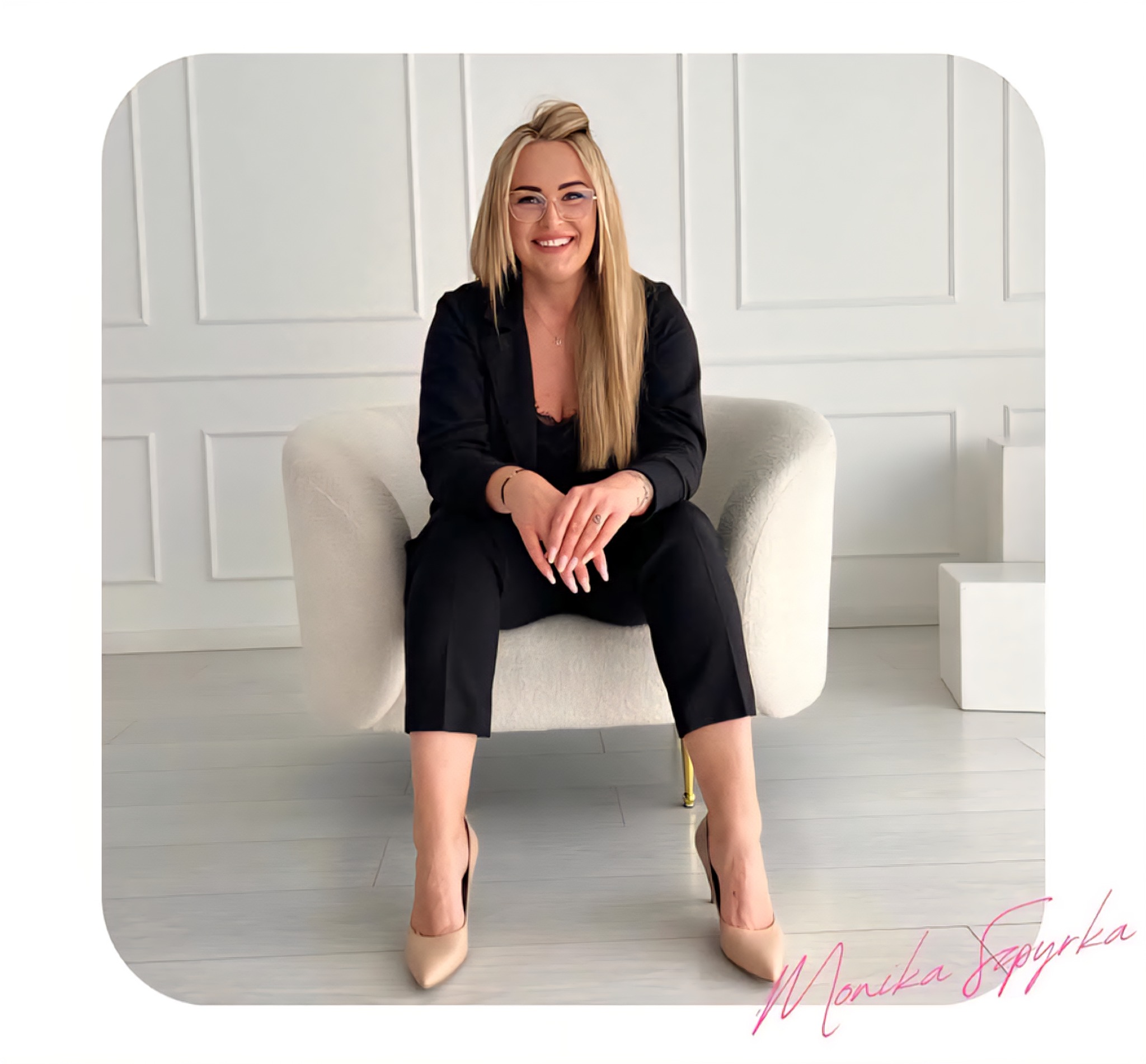 Uśmiechnięta blondynka w okularach, czarnym garniturze i beżowych szpilkach siedzi w białym fotelu na tle białej ściany z panelami. Sygnatura w prawym dolnym rogu.