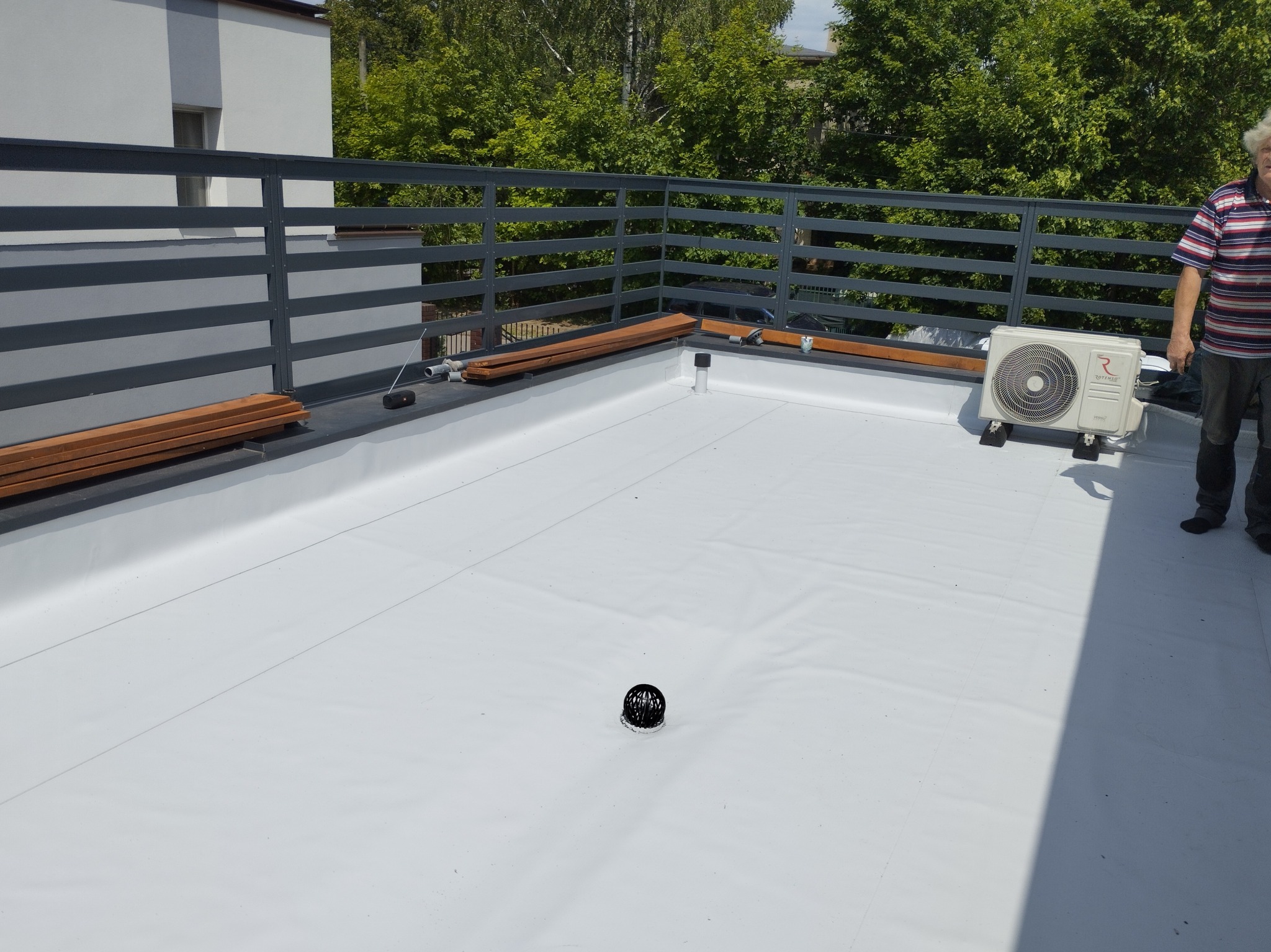 Biały dach płaski z kratą, drewnianymi elementami i klimatyzatorem. Widoczny odpływ wody. Osoba w tle.