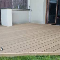 Nowoczesny taras kompozytowy w odcieniach beżu, z widoczną strukturą drewna, przylegający do budynku z otwartymi drzwiami. Tło stanowi drewniany płot i zieleń trawnika.