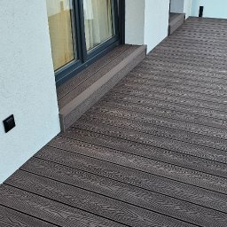 Drewniany Akcent Stolarstwo - Nowoczesny taras kompozytowy z desek w odcieniach brązu, przylegający do budynku z dużym oknem i białymi ścianami. Widoczny fragment balustrady.