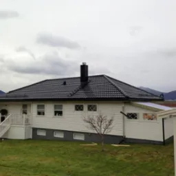 Dom z czarnym dachem z dachówki, białą elewacją i schodami wejściowymi. W tle góry i pochmurne niebo. Widoczne detale wykończenia i otoczenie domu.