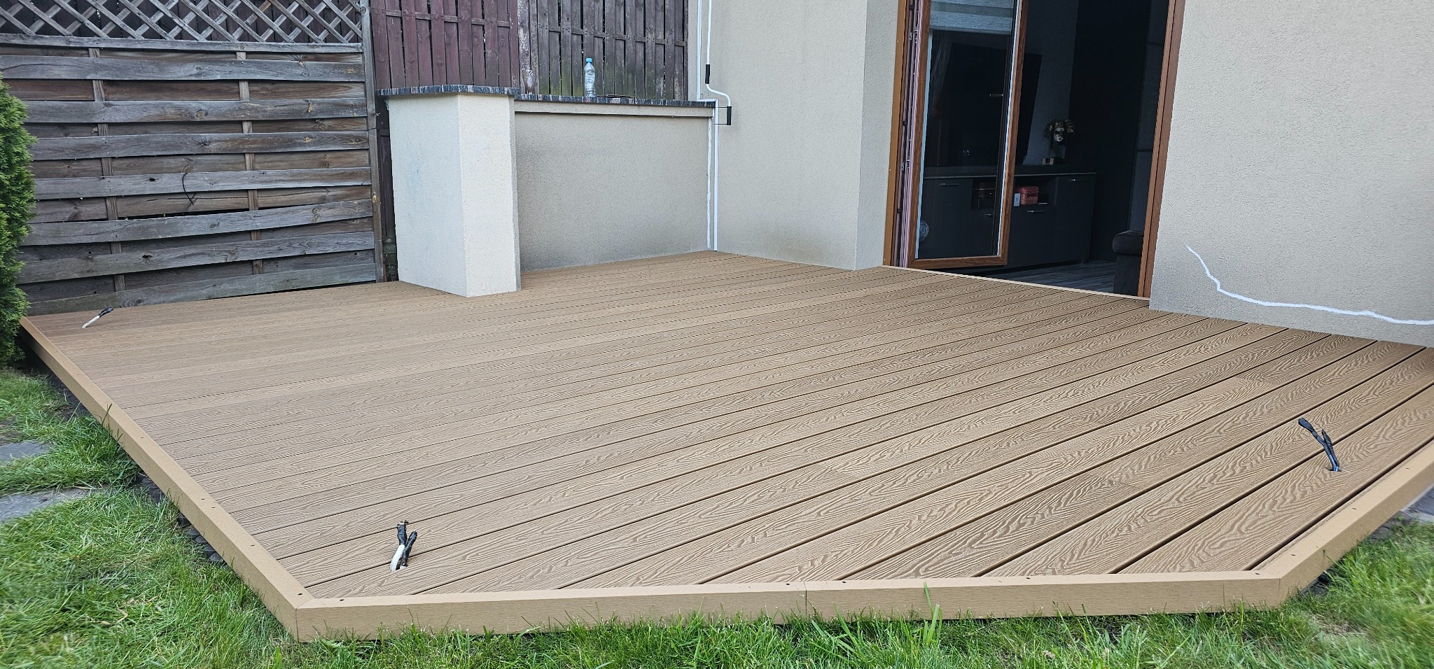 Nowoczesny taras kompozytowy w odcieniach beżu, z widoczną strukturą drewna, przylegający do budynku z otwartymi drzwiami. Tło stanowi drewniany płot i zieleń trawnika.