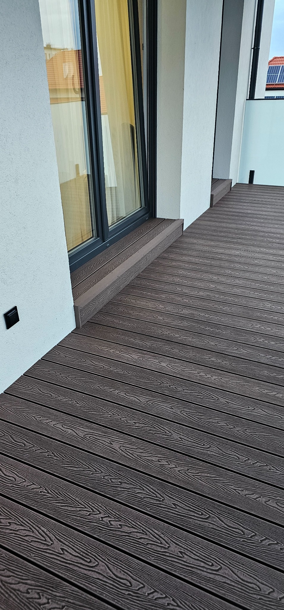 Nowoczesny taras kompozytowy z desek w odcieniach brązu, przylegający do budynku z dużym oknem i białymi ścianami. Widoczny fragment balustrady.
