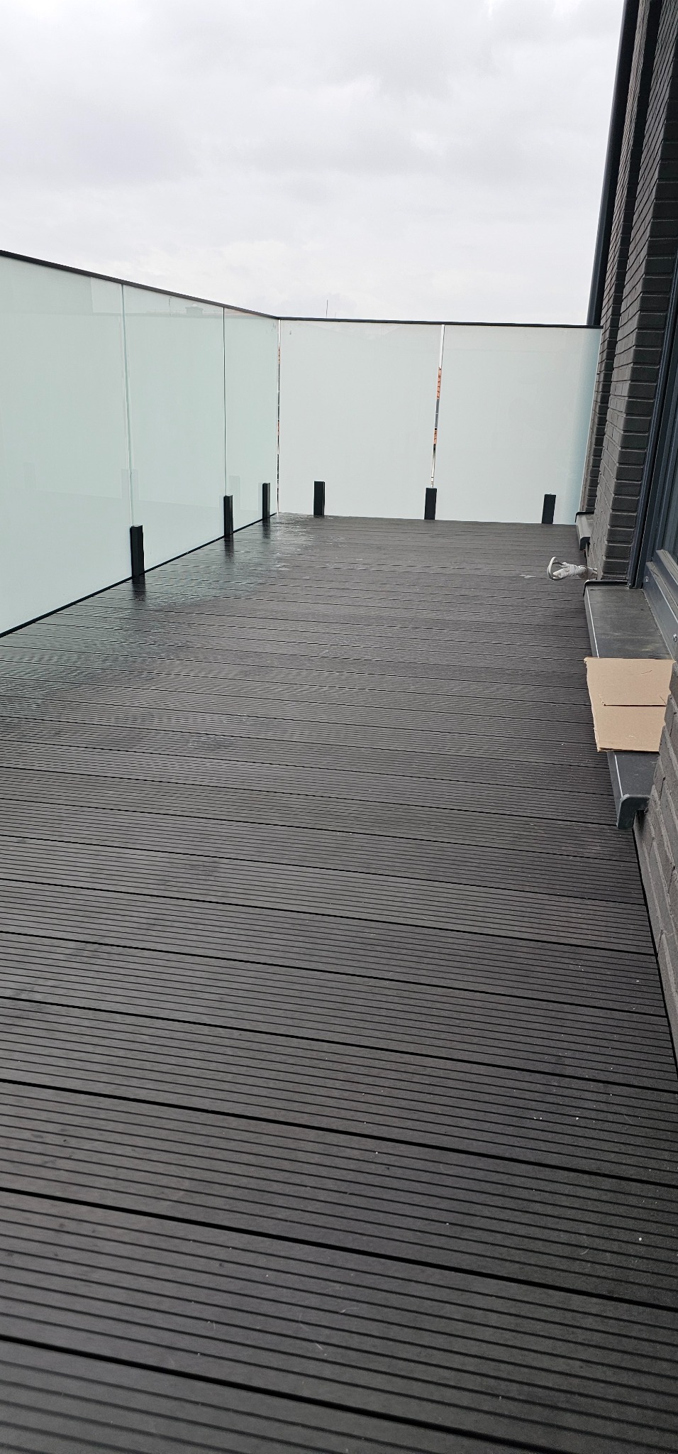 Nowoczesny taras z ciemnych desek kompozytowych, zabezpieczony szklaną balustradą. Widok na pochmurne niebo.