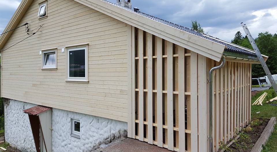 Drewniana konstrukcja budynku z elementami ażurowymi, widoczna drabina i rynna. Elewacja w jasnym odcieniu drewna, częściowo wykończona deskami.