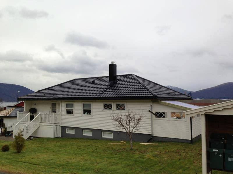 Dom z czarnym dachem z dachówki, białą elewacją i schodami wejściowymi. W tle góry i pochmurne niebo. Widoczne detale wykończenia i otoczenie domu.