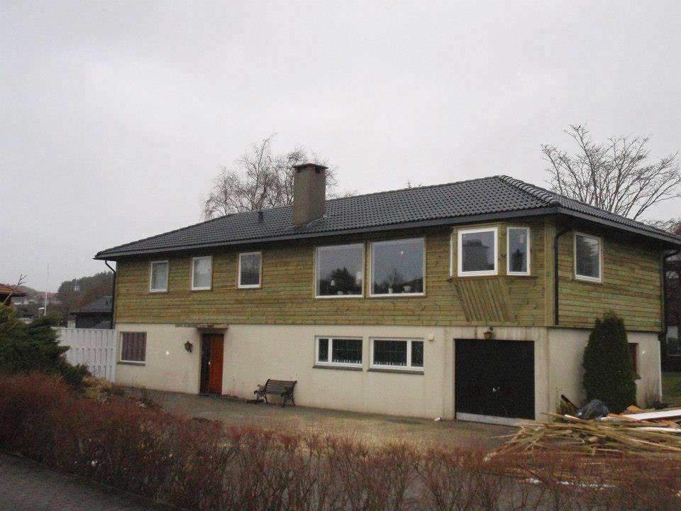 Dwukondygnacyjny dom z nowym, ciemnym dachem i świeżo obłożoną drewnianą elewacją. Widoczne okna, drzwi i stos drewna przy garażu. Pochmurne niebo.