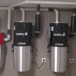 Instalacja centralnego odkurzania Husky: czerwony Hurricane 660 i dwa czarne Titan, rury PCV pod sufitem, szara ściana w płytkach, gniazdko elektryczne obok.