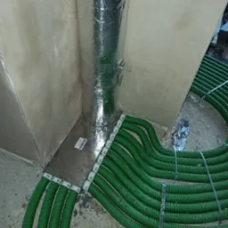 Fragment instalacji rekuperacji: srebrna rura w folii aluminiowej i wiązka zielonych rur wentylacyjnych na podłodze w rogu pomieszczenia. Etap montażu instalacji.