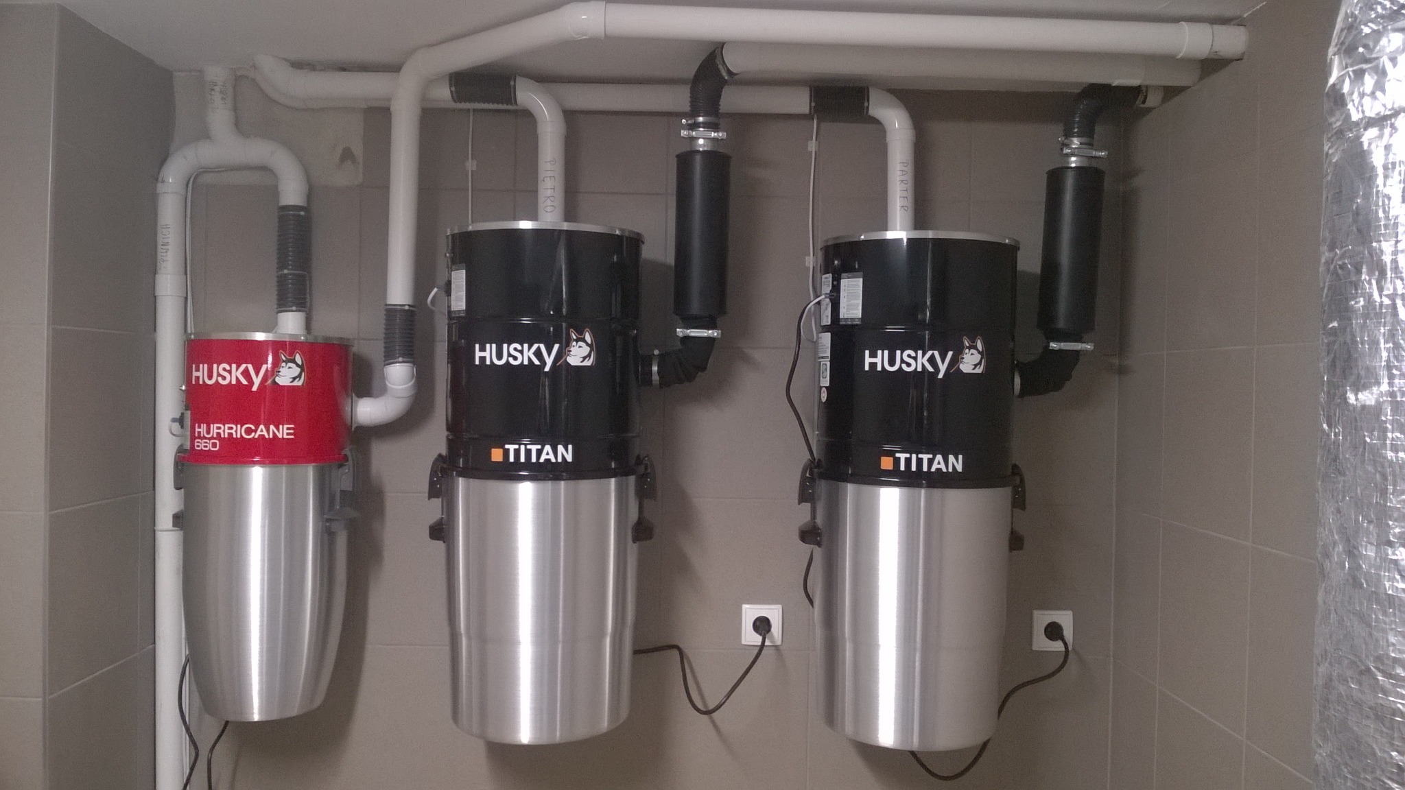 Instalacja centralnego odkurzania Husky: czerwony Hurricane 660 i dwa czarne Titan, rury PCV pod sufitem, szara ściana w płytkach, gniazdko elektryczne obok.