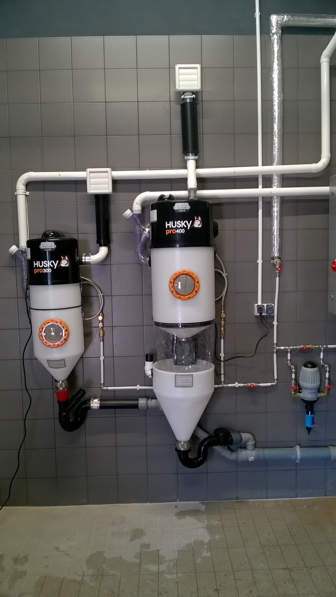 Dwa urządzenia Husky Pro 300 i 400 zamontowane na szarej, kafelkowej ścianie z widocznymi rurami i przewodami wentylacyjnymi. Instalacja w pomieszczeniu z szarą podłogą.