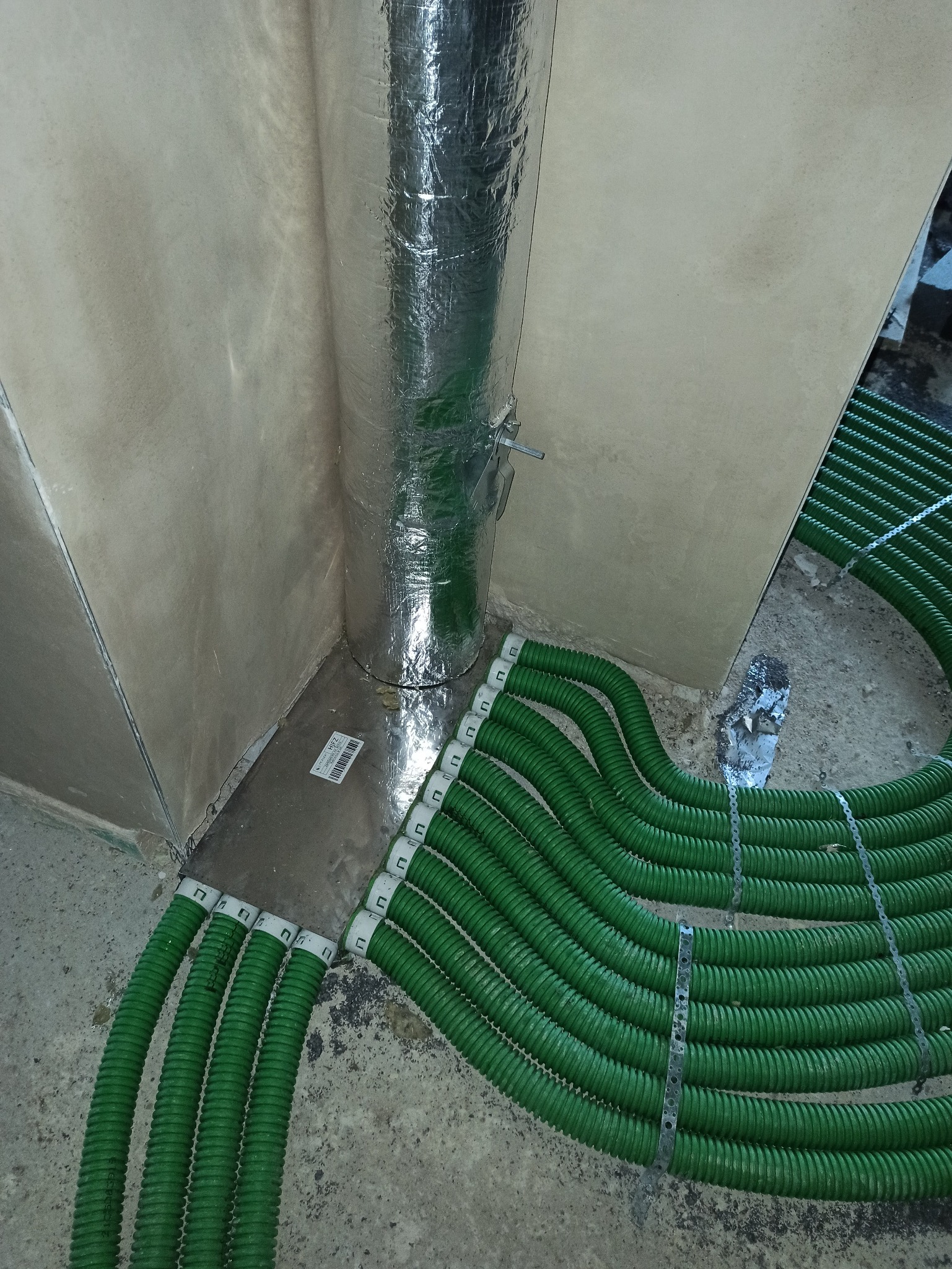 Fragment instalacji rekuperacji: srebrna rura w folii aluminiowej i wiązka zielonych rur wentylacyjnych na podłodze w rogu pomieszczenia. Etap montażu instalacji.