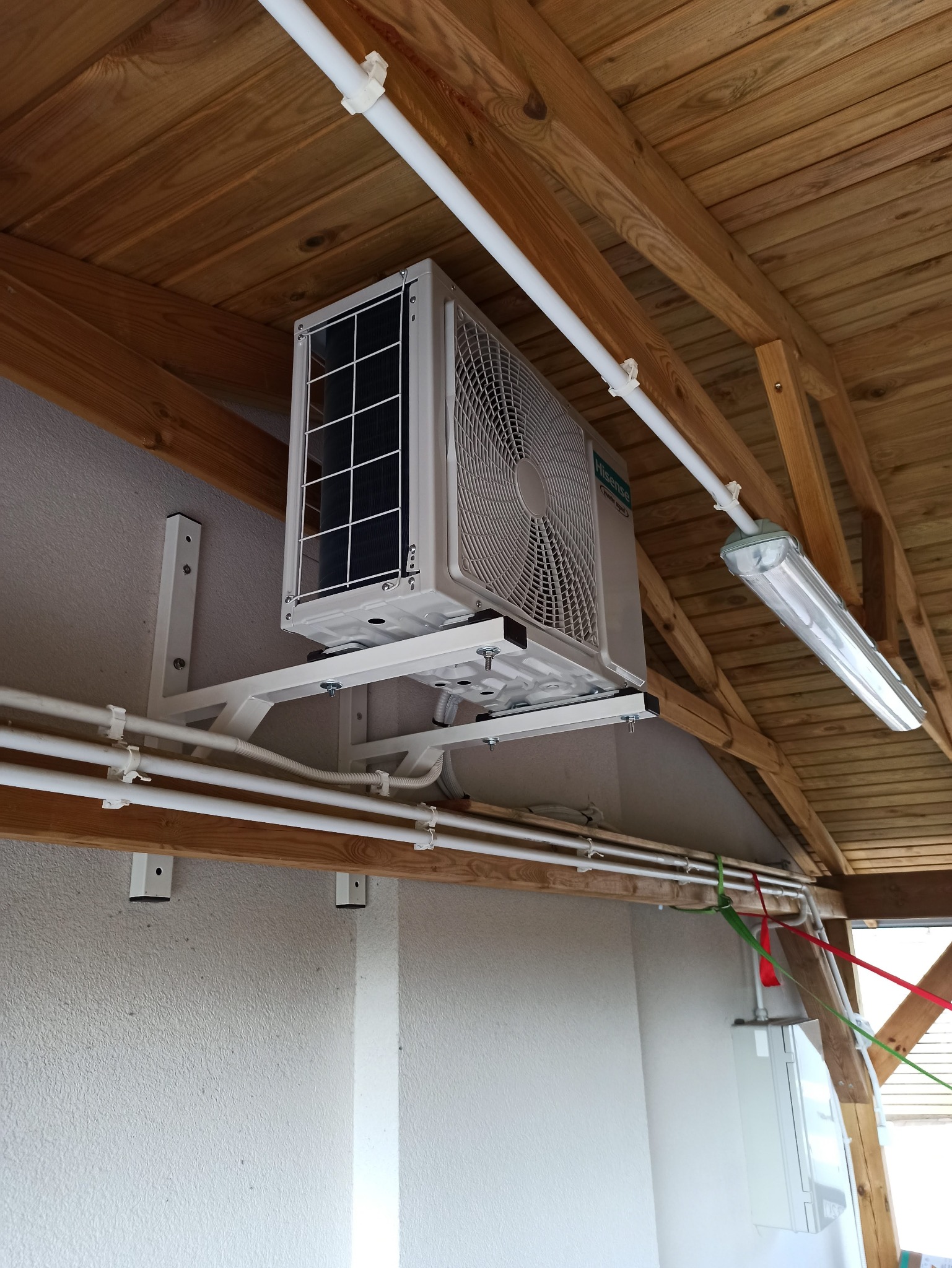 Klimatyzator Hisense zamontowany na białym wsporniku pod drewnianym stropem, widoczne białe rurki instalacyjne i oświetlenie. Ujęcie z dołu, podkreślające precyzję montażu.