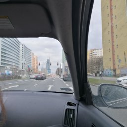 Michał Stolarczyk - Widok z wnętrza samochodu na ulicę miejską z wysokimi budynkami i wieżowcami w oddali, pochmurne niebo. Perspektywa zza deski rozdzielczej auta.