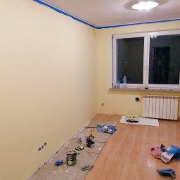 Michał Stolarczyk - Pokój po malowaniu ścian na żółto, z niebieską taśmą malarską przy suficie. Widoczne farby, wałki i zabezpieczona podłoga. Okno z grzejnikiem.