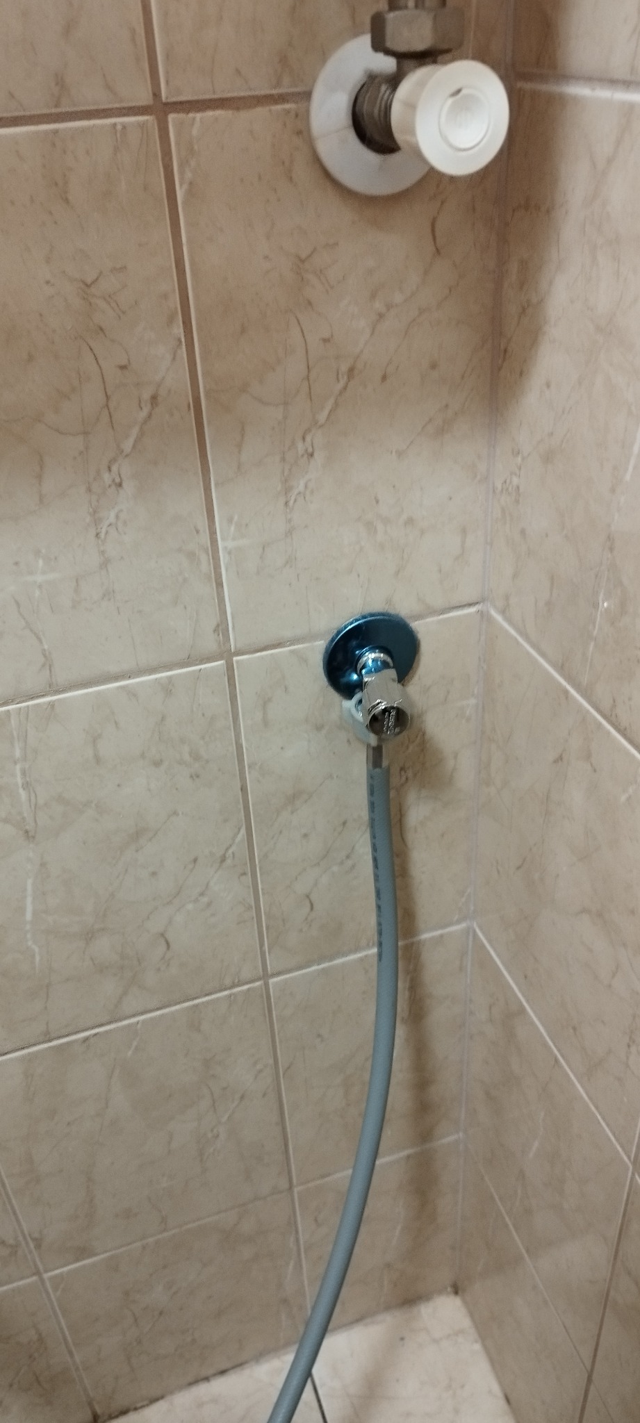 Błyszczący zawór kątowy z podłączonym szarym wężykiem na tle beżowych, marmurkowych płytek ściennych. Drugi zawór widoczny w górnym rogu.