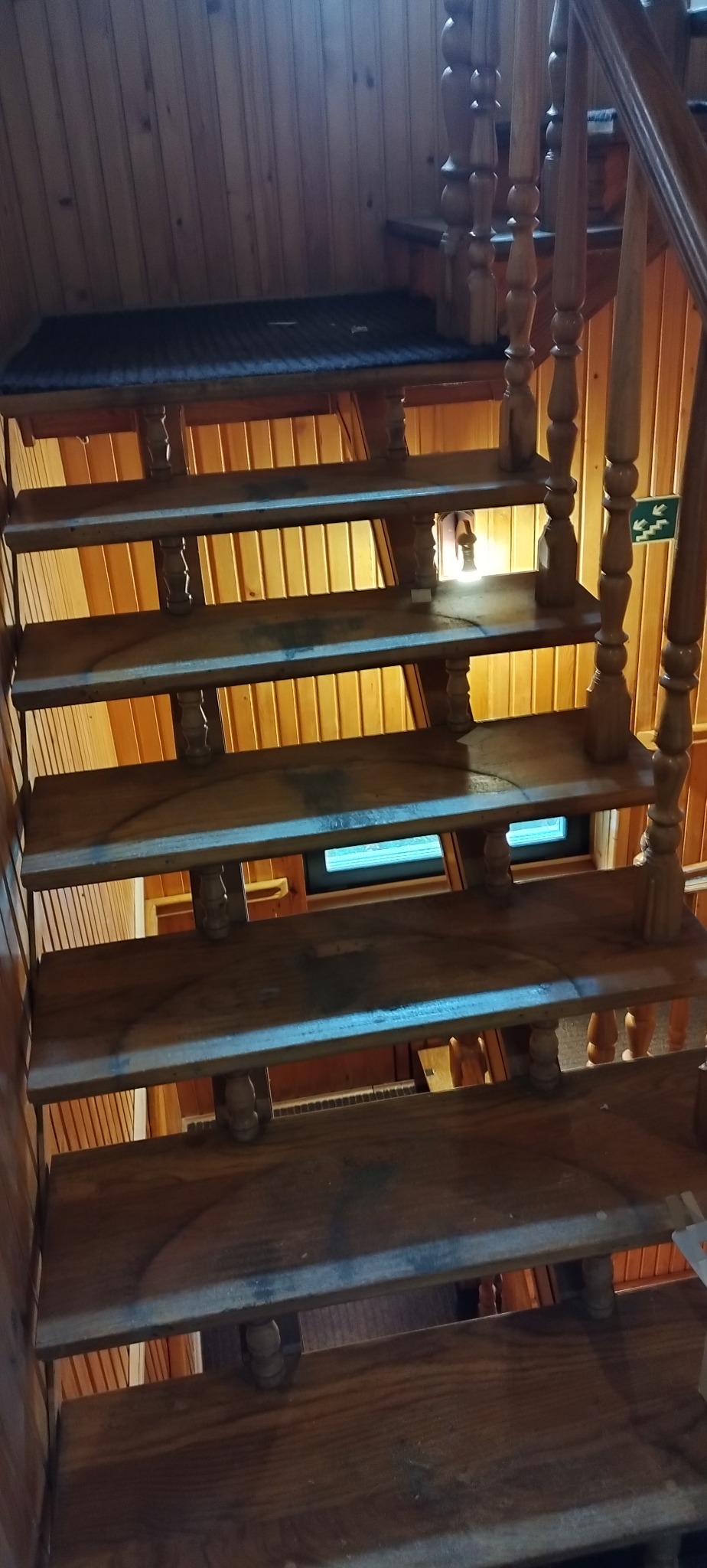 Drewniane schody z toczonymi balustradami w domu z boazerią; widok z dołu, na stopniach widoczne ślady użytkowania, w tle okno i drewniane ściany.