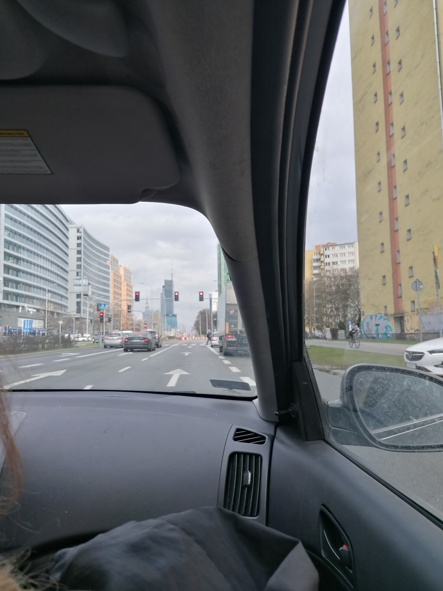 Widok z wnętrza samochodu na ulicę miejską z wysokimi budynkami i wieżowcami w oddali, pochmurne niebo. Perspektywa zza deski rozdzielczej auta.