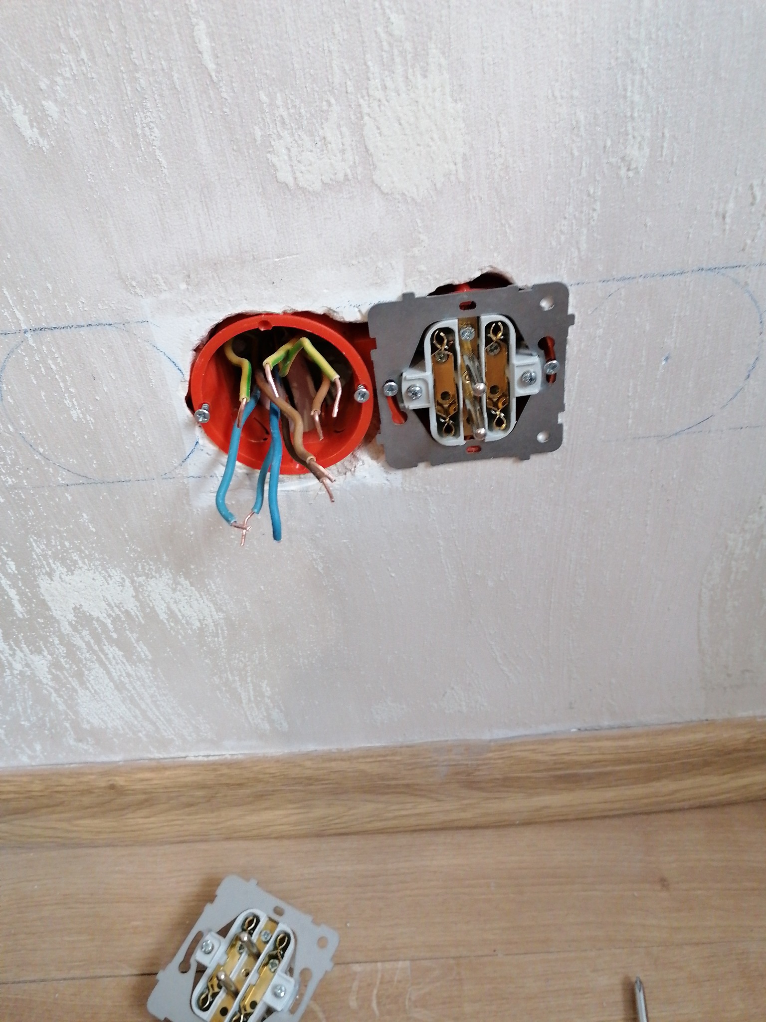 Gniazdko elektryczne w trakcie montażu: puszka z przewodami i częściowo zamontowany mechanizm na tle pomalowanej ściany i drewnianej listwy przypodłogowej.