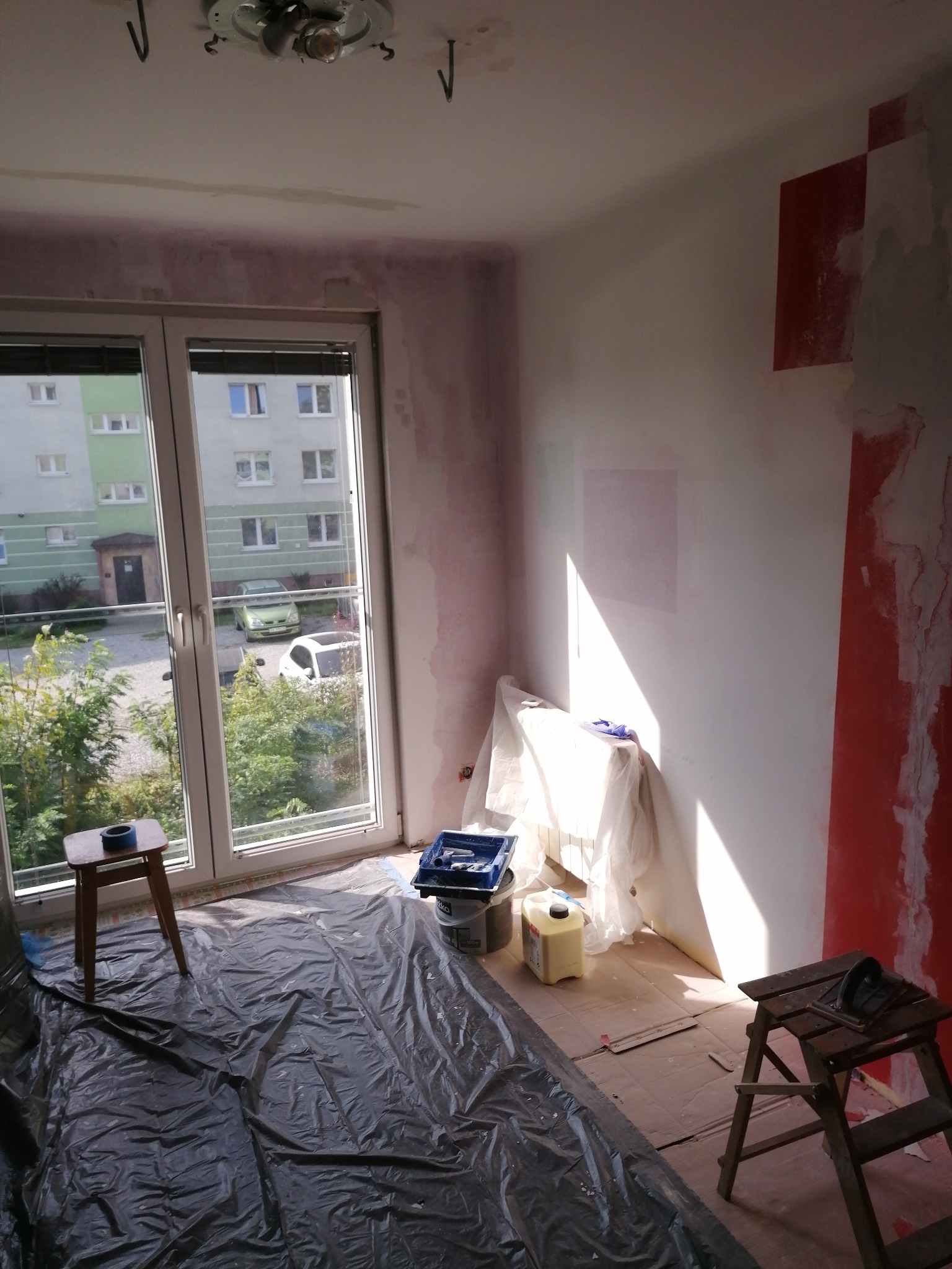 Pokój w trakcie malowania: zabezpieczona folią podłoga, drabina malarska, wiadro z farbą. Widoczne ślady po taśmie malarskiej na ścianach.