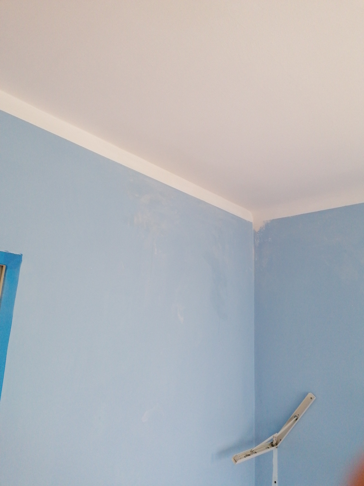Świeżo pomalowany na błękitno róg pokoju z białym sufitem i listwą. Na ścianie widoczne ślady po wałku malarskim i rozłożona suszarka na pranie.
