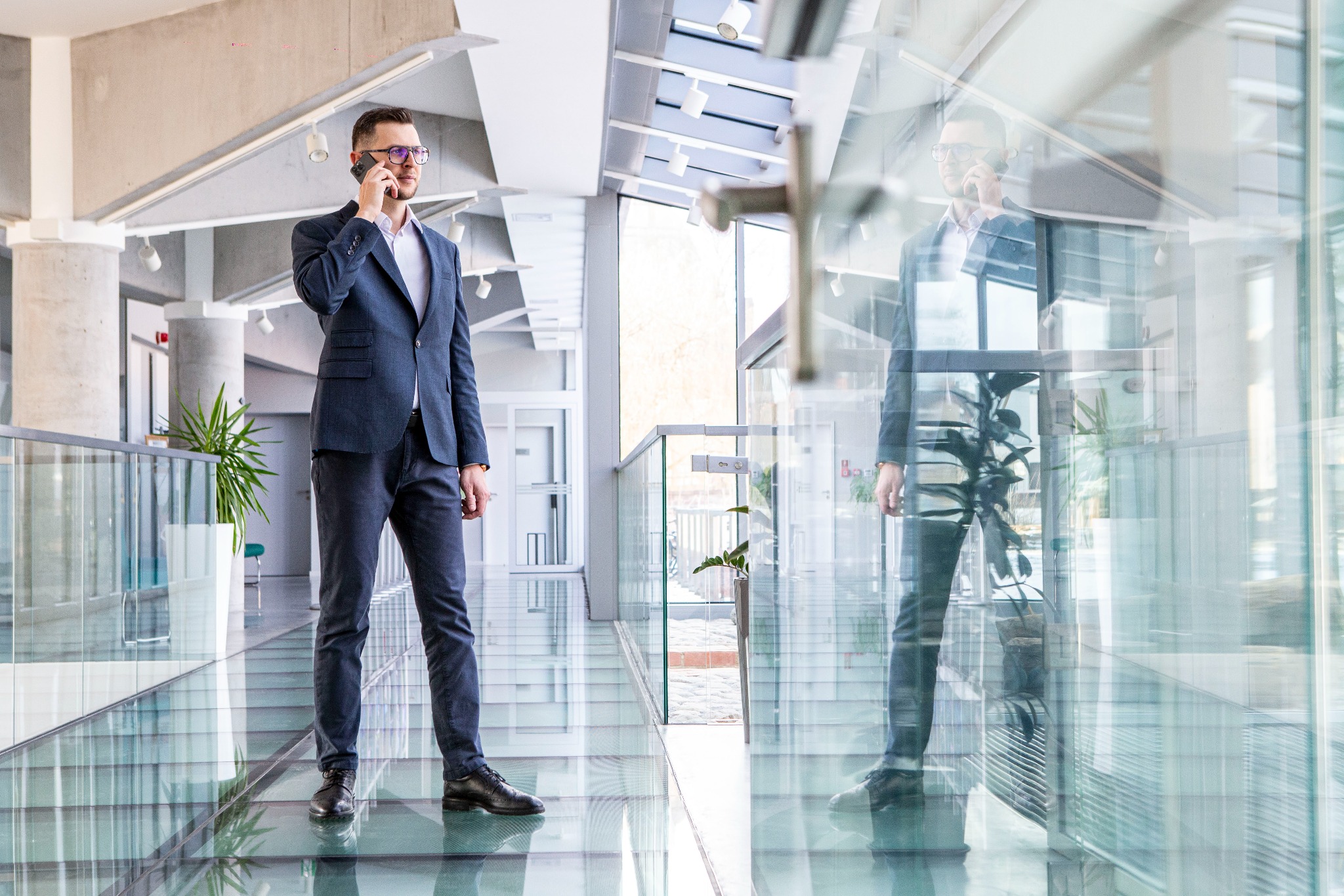 Biznesmen w garniturze rozmawia przez telefon w nowoczesnym biurze z przeszkloną podłogą, odbijającą jego postać i otoczenie. Widoczne rośliny i jasne wnętrze.