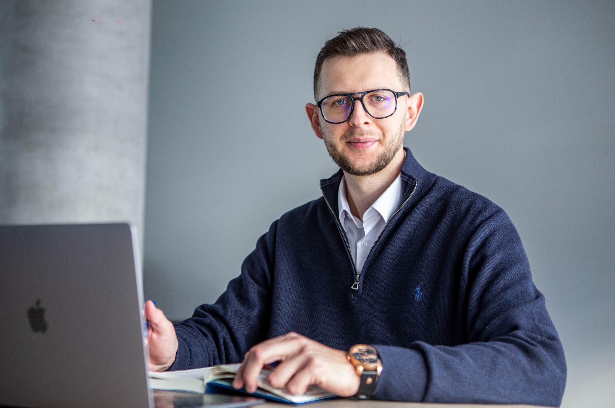 Mężczyzna w okularach siedzi przy biurku z laptopem i notatnikiem, uśmiecha się, ubrany w granatowy sweter i białą koszulę. Stonowane tło, naturalne światło.